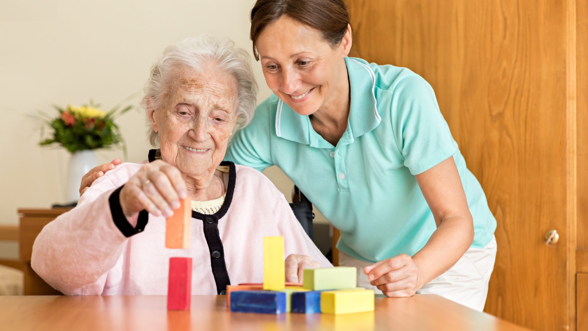 Elderly woman with dementia smiling while playing with colorful blocks, supported by a compassionate carer—illustrating the warmth and engagement provided through quality dementia care at home.