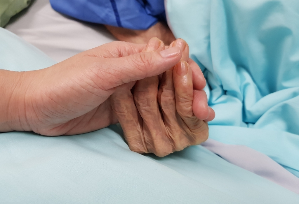 A compassionate moment during end of life care at home, showing a carer gently holding the hand of an elderly patient resting in bed