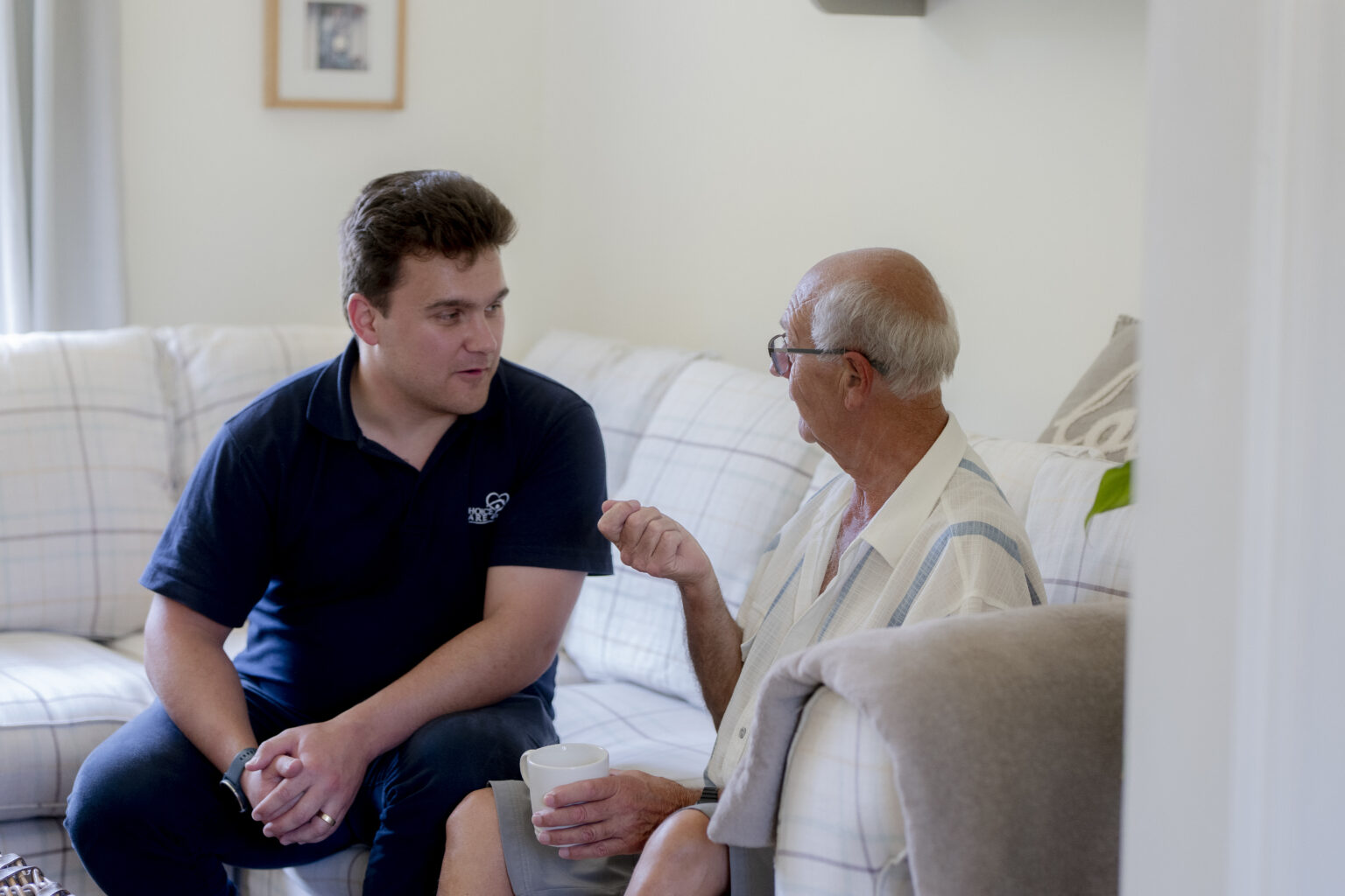 A carer talking with an elderly man in his living room, illustrating the personal support differences between live-in care vs. visiting care.