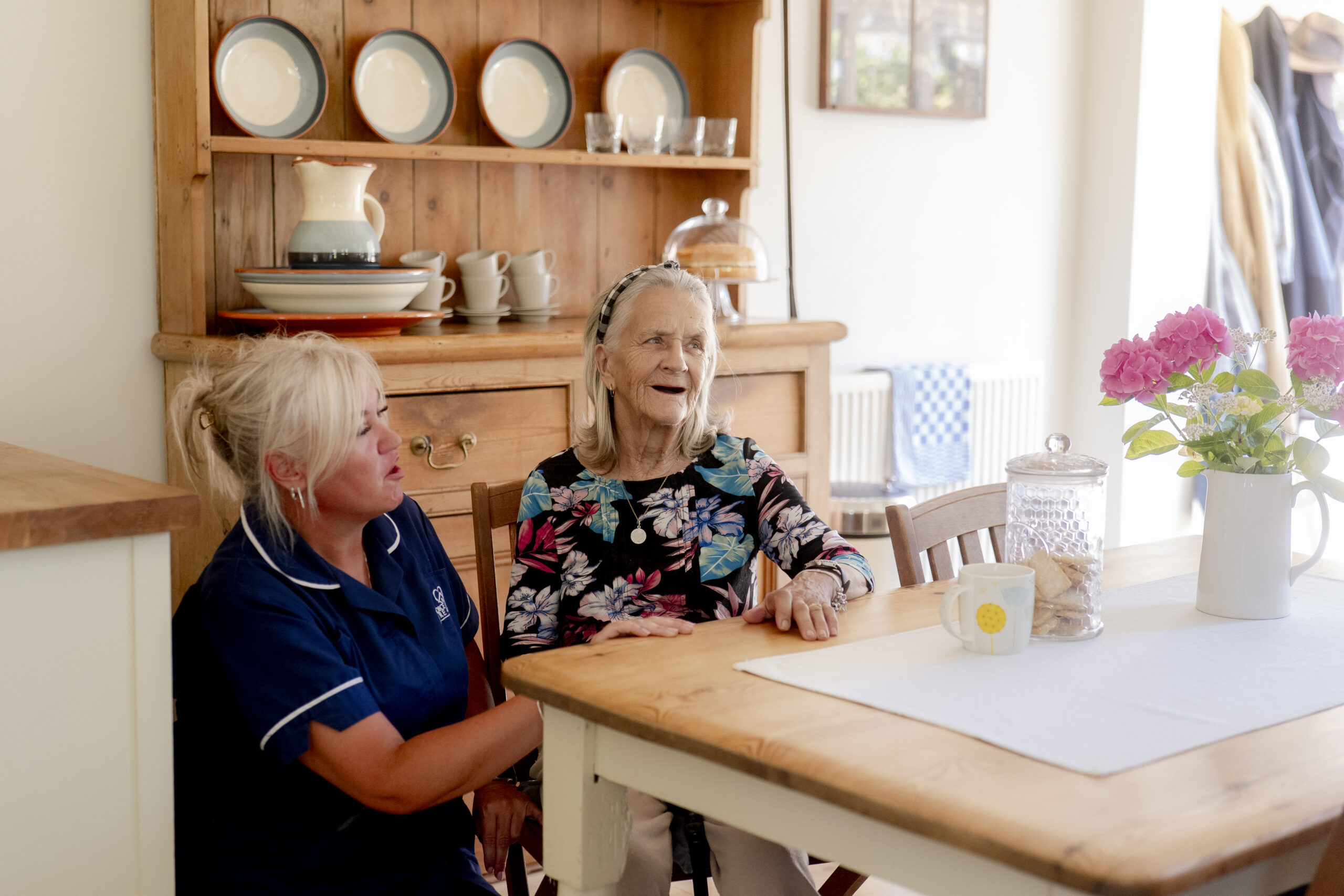 Carer chatting with an elderly woman in the kitchen, illustrating the benefits of private home care for seniors through companionship and social interaction.