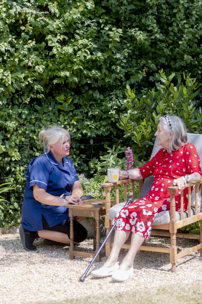 Carer providing in-home care services for seniors, talking warmly to an elderly person sitting in a chair and holding a glass.