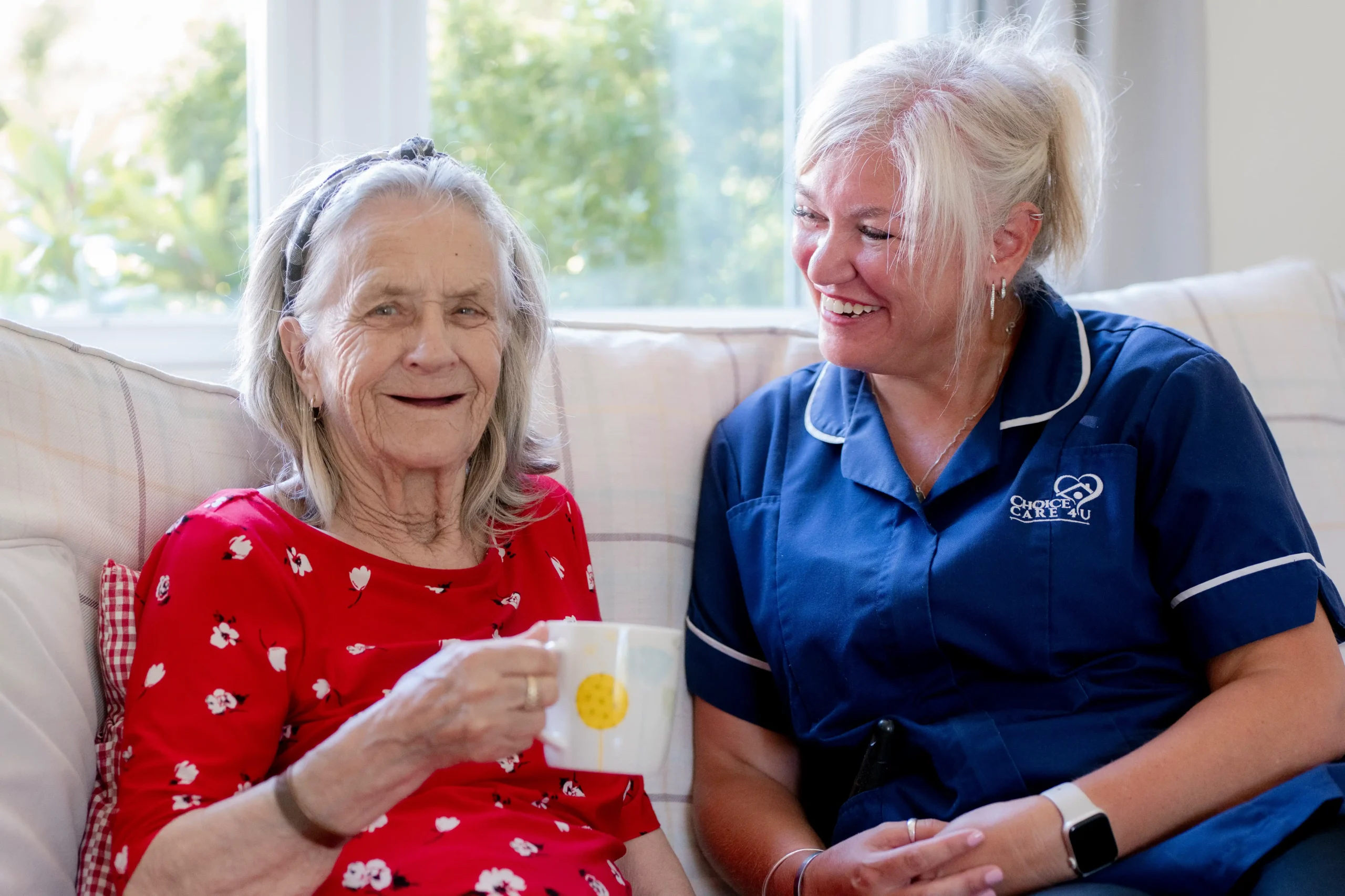 Carer from Choice Care 4U smiling warmly at an older client during a relaxed moment; both are seated, and the client is holding a cup, a comforting scene showcasing quality care at home services.