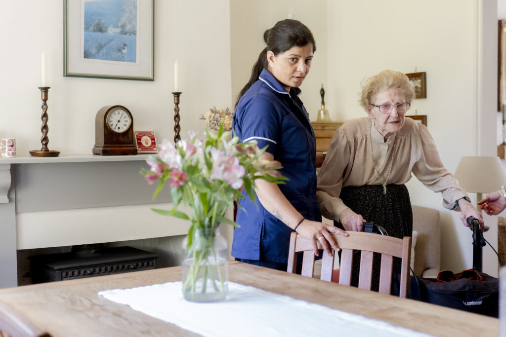 A friendly carer from Choice Care 4U supporting an elderly woman as they walk together inside her home, representing compassionate help at home care services.
