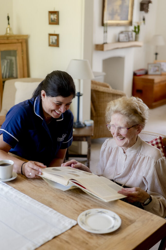 Carer from Choice Care 4U providing companionship care while reading a book and smiling with an elderly woman at a dining table.