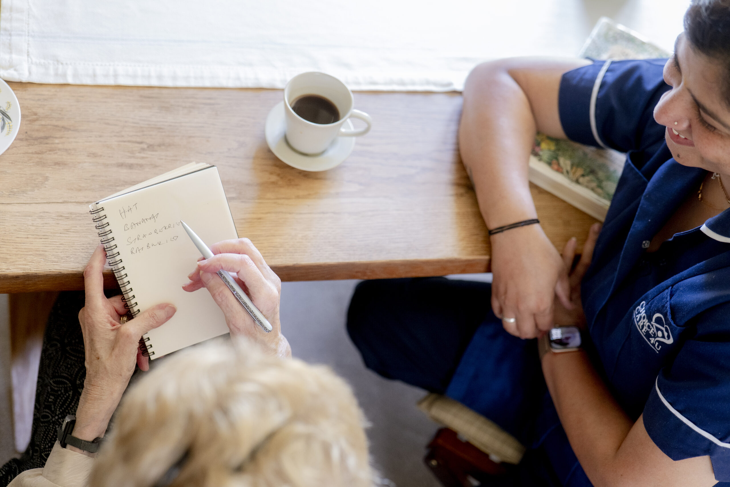 Carer supporting an older woman with a pen and notebook, offering compassionate dementia and palliative care at home.
