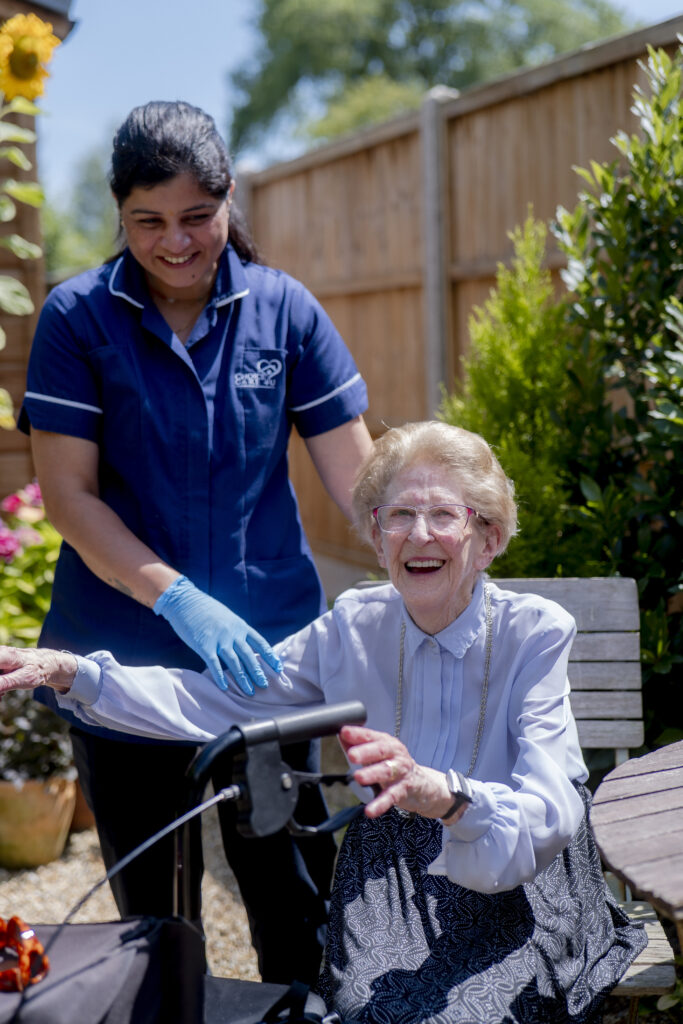 A friendly carer from Choice Care 4U standing beside an older woman smiling warmly in a chair, representing quality companionship care and emotional support for the elderly.